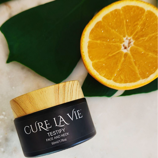 A jar of Cure La Vie's TESTIFY face cream with a wooden lid, displayed next to a sliced orange and green leaf on a white surface.