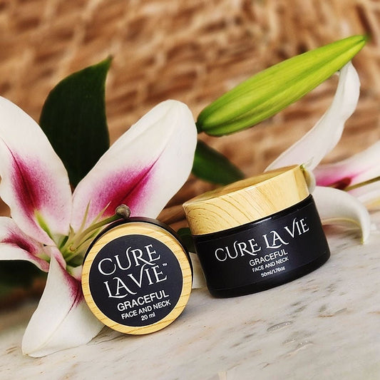 Two jars of Cure La Vie skin care products, both labeled 'GRACEFUL' for face and neck, one full size, one travel size, placed on a surface with a flower in the background.