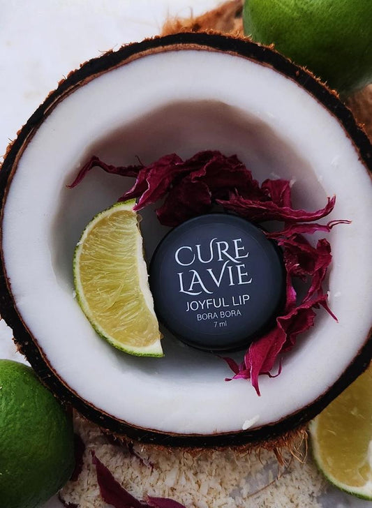 A Cure La Vie "JOYFUL LIP" lip balm, "BORA BORA", sitting next to a cut in half lime and flower petals inside a coconut, with whole limes around the coconut.