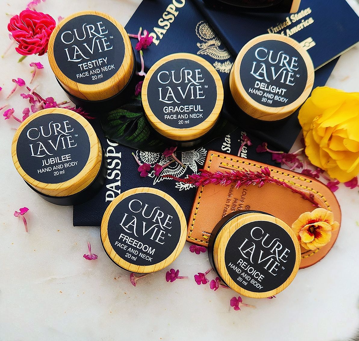 Six small travel jars of skin care cream labeled 'CURE LA VIE' with the names TESTIFY, GRACEFUL, DELIGHT, JUBILEE, FREEDOM, and REJOICE, placed on a white marble surface with travel items and flowers surrounding.